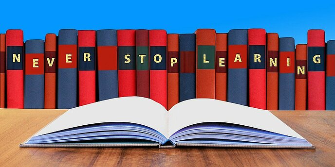 Man sieht eine Reihe Bücher in verschiedenen Farben und davor ein aufgeschlagenes Buch. Auf der Bücherreihe steht in englischer Sprache "Never stop Learning"