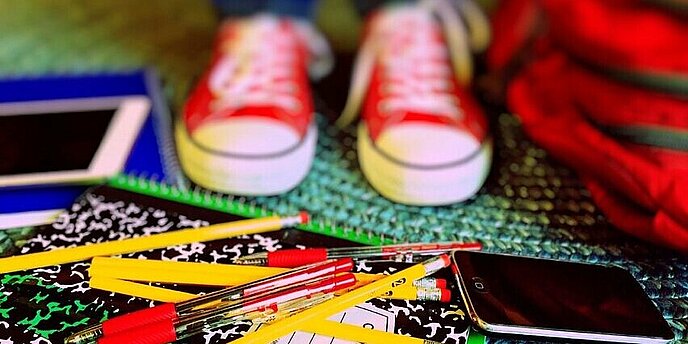 Rote Turnschuhe, rote Kugelschreiber, Bleistifte, ein Handy und ein Tablet sind zu sehen