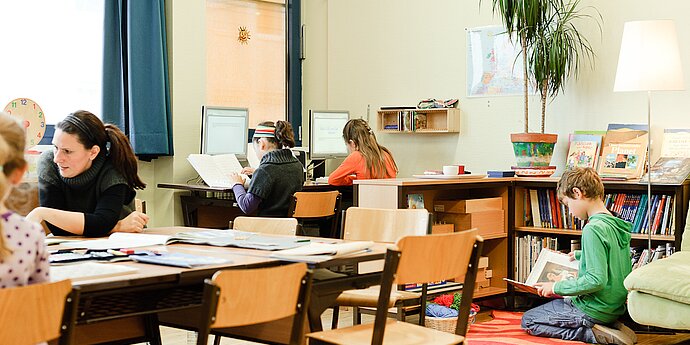 Eine Lehrerin in einem Klassenzimmer mit mehreren Schülerinnen und Schülern im Unterricht. Zwei Mädchen sitzen beim Computer, ein Bub liest in einem Buch.